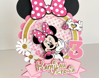Puede incluir: Topper de pastel de Minnie Mouse rosa y dorado con fondo brillante, una margarita blanca y el nombre "Kamilah Victoria" en una pancarta rosa. El número "3" está en el lado derecho del topper.