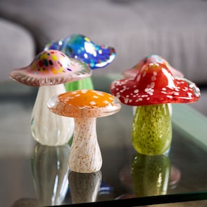 May include: Five glass mushroom figurines in various colors, including pink, blue, red, green, and orange. Each mushroom has white polka dots on the top and a textured stem.