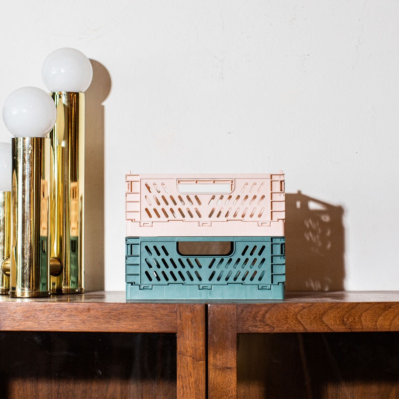 Puede incluir: Dos cajas de almacenamiento de pl&aacute;stico apilables, una rosa y otra verde azulado, con un patr&oacute;n de rejilla. Las cajas est&aacute;n sobre una superficie de madera.