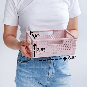 Puede incluir: Un contenedor de almacenamiento plegable rosa con asa. El contenedor mide 15,2 cm de alto, 8,9 cm de ancho y 21,6 cm de largo.