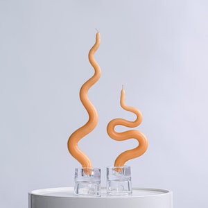 May include: Two orange wax candles in glass holders. The candles are shaped like a wavy line. The candles are on a white surface.