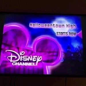 Può includere: Uno schermo televisivo che mostra il logo viola di Disney Channel con un contorno luminoso. Il testo Halloweentown High starts now è visibile in bianco su uno sfondo blu scuro con una luna piena e la sagoma di un edificio.