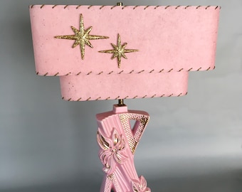 May include: A pink ceramic table lamp with a pink fabric lampshade. The lampshade has gold glitter stars and a stitched edge. The base of the lamp is shaped like a stylized flower.