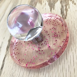 May include: A clear glass knob with a silver base sits on a pink glitter-filled resin base.