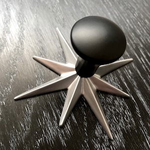 May include: A silver star-shaped base with eight points and a black knob on top. The knob is smooth and round.