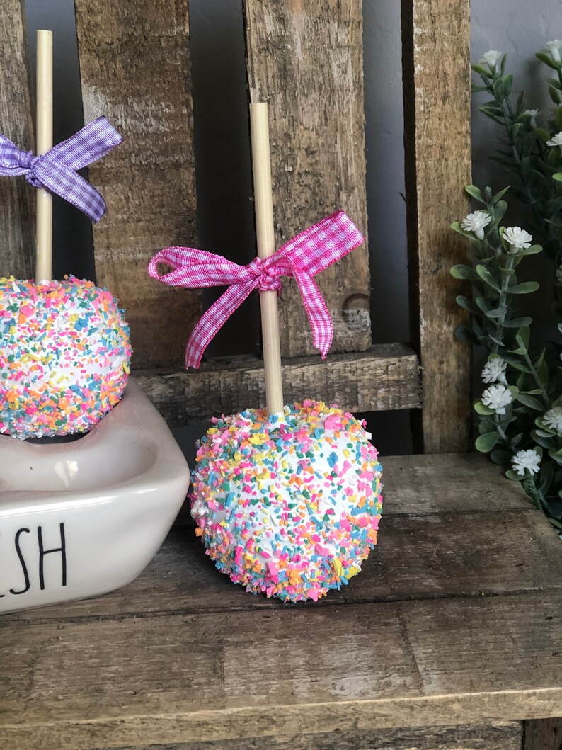 May include: Two white chocolate apples covered in colorful sprinkles on wooden sticks with pink and purple gingham bows. The apples are sitting on a white ceramic dish with the word "Dish" written on it.