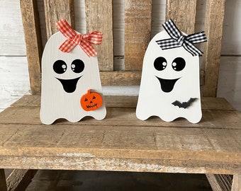 Può includere: Due ornamenti di fantasmi in legno bianco con occhi e bocche neri. Un fantasma ha un fiocco a scacchi rosso e bianco e una piccola zucca arancione con un viso di zucca. L'altro fantasma ha un fiocco a scacchi bianco e nero e un piccolo pipistrello nero.
