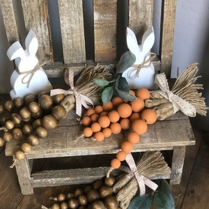 May include: Three wooden bead garlands with different coloured beads, tied with twine and a ribbon, and placed on a wooden chair with two white wooden bunny figurines.
