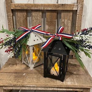 Op de afbeelding: Twee decoratieve lantaarns met rode, witte en blauwe lintaccenten. De lantaarns staan op een houten oppervlak en zijn versierd met een boeket witte en blauwe bloemen.