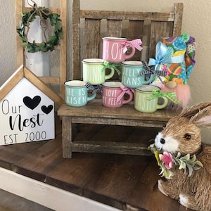 May include: A set of six pastel-colored ceramic mugs with Easter-themed sayings. The mugs are stacked on a wooden bench with a bunny rabbit figurine and a sign that reads "Our Nest EST 2000".