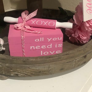 May include: Pink wooden blocks stacked on top of each other with the words "all you need is love" written on them. A pink rolling pin with a white handle and the word "XOXO" written on it is tied with a pink and white gingham ribbon on top of the blocks.