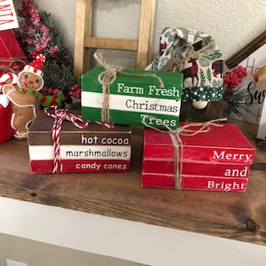 Może przedstawiać: Trzy drewniane klocki ułożone jeden na drugim, ozdobione świątecznymi powiedzeniami. Górny klocek jest czerwony i mówi "Wesołych i jasnych świąt". Środkowy klocek jest czerwony i mówi "gorąca czekolada pianki cukierki trzcinowe". Dolny klocek jest zielony i mówi "Świeże choinki z farmy".