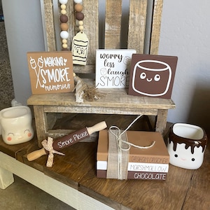 May include: A rustic wooden chair with a brown and white beaded garland, three wooden signs with the text "Making S'more Memories", "Worry Less Laugh S'more", and a cartoon marshmallow, a brown and white rolling pin with the text "S'more Please", a stack of three books with the text "Graham Cracker, Marshmallow, Chocolate", and two white ceramic mugs with cartoon marshmallow faces.