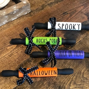 May include: Four mini rolling pins decorated with Halloween themes. The rolling pins are painted in different colours, including green, white, purple, and orange. Each rolling pin has a black ribbon tied around it and features a different Halloween phrase: "Spooky", "Hocus Pocus", "Trick or Treat", and "Halloween".