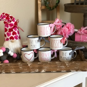 May include: A collection of white mugs with black rims and handles, each inscribed with a Valentine's Day message in pink or black. The mugs are arranged on a wooden tray, alongside a gnome and decorative beads. Ribbons adorn the mugs.