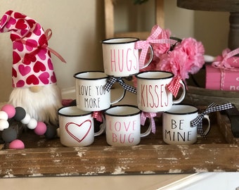 May include: A collection of white mugs with black rims and handles, each inscribed with a Valentine's Day message in pink or black. The mugs are arranged on a wooden tray, alongside a gnome and decorative beads. Ribbons adorn the mugs.