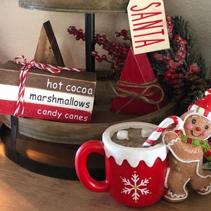 Puede incluir: Una bandeja de madera de dos niveles decorada para Navidad. El nivel superior tiene una tela de cuadros rojos y negros, cuerda blanca y una etiqueta de madera que dice "SANTA". El nivel inferior tiene un letrero de madera que dice "hot cocoa marshmallows candy canes", una taza roja y blanca con un diseño de copo de nieve y un hombre de jengibre sosteniendo un bastón de caramelo.