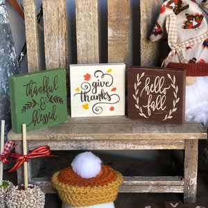 Pode incluir: Três placas de madeira com dizeres temáticos de outono: "thankful & blessed", "give thanks" e "hello fall". As placas são exibidas em um banco de madeira rústico com um gnomo e outras decorações de outono.