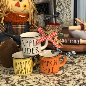 May include: Three ceramic mugs with fall-themed designs. The mugs are white with black lettering and a black rim. The mugs are labeled "Apple Cider", "Harvest", and "Pumpkin Spice".