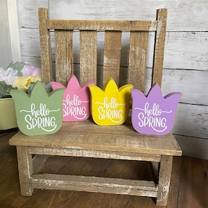May include: Four wooden tulip-shaped signs with the text "hello spring" in white lettering. The signs are painted in different colors: green, pink, yellow, and purple. The signs are arranged on a rustic wooden bench.