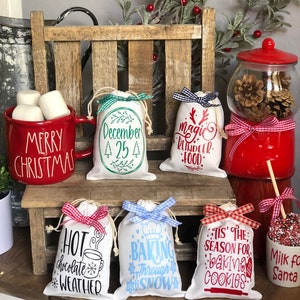 Puede incluir: Seis pequeñas bolsas de regalo de tela blanca con lazos de cinta de vichy rojo y blanco. Cada bolsa tiene un diseño y un texto diferentes con temática navideña, que incluyen "Diciembre 25", "Comida mágica para renos", "Clima de chocolate caliente", "Hornear a través de la nieve" y "Es la temporada para hornear galletas".