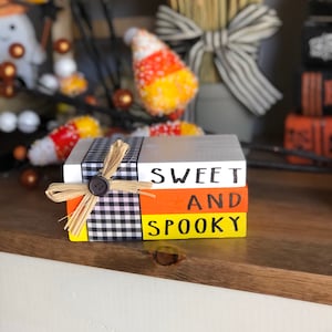 Può includere: Tre blocchi di legno impilati con un motivo a scacchi bianco e nero, strisce arancioni e gialle e le parole "Sweet and Spooky" in lettere nere.