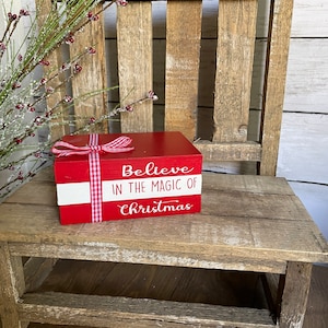 Puede incluir: Una caja de madera roja con una franja blanca y una cinta de cuadros rojos y blancos. La caja tiene las palabras "Believe in the magic of Christmas" escritas en ella.