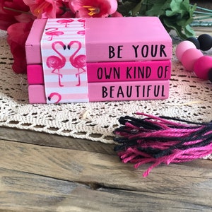 May include: Three pink stacked books with the words "BE YOUR OWN KIND OF BEAUTIFUL" on the front. The books are decorated with pink flamingos and white stripes.