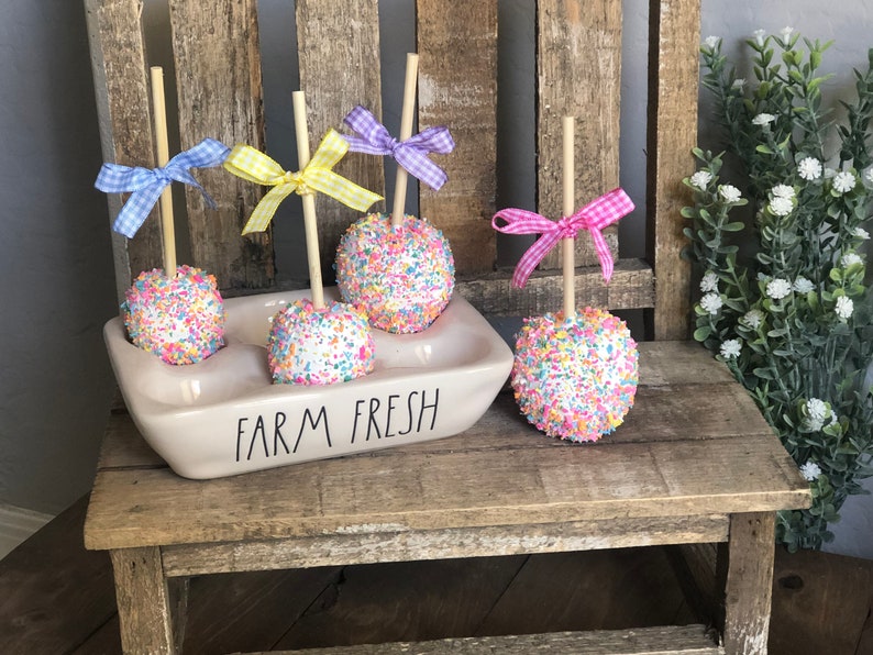 May include: Four white cake pops decorated with colorful sprinkles, each with a wooden stick and a pastel colored ribbon bow. The cake pops are displayed in a white ceramic holder with the words "FARM FRESH" printed on the front.