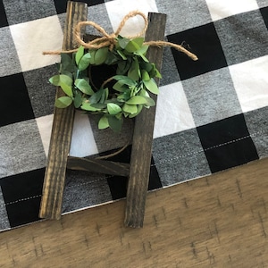 May include: A small wooden ladder with a green leafy wreath tied to the top rung. The ladder is sitting on a black and white checked tablecloth.