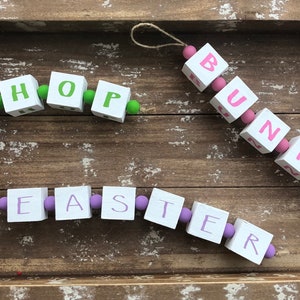 May include: Three wooden bead garlands with the words "Easter", "Hope", and "Bunny" spelled out in pink and purple paint. The garlands are hanging on a wooden tray.