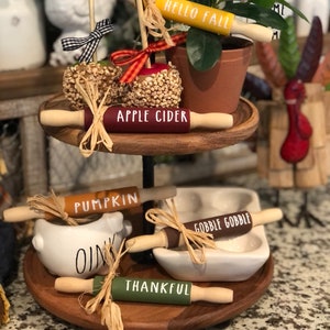 May include: A tiered wooden tray featuring a selection of miniature rolling pins with autumnal themes. The rolling pins are decorated with phrases such as "Hello Fall", "Apple Cider", "Pumpkin", "Gobble Gobble", "Thankful", and "Harvest".