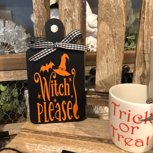 Könnte beinhalten: Schwarzes Holzschild mit einem Hexenhut- und Fledermausdesign. Das Schild sagt "Witch Please" in orangefarbenem Text mit einem schwarzen Besen und Sternen. Das Schild ist mit einem schwarz-weiß karierten Band gebunden.