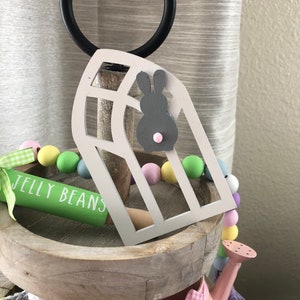 May include: A tiered wooden tray decorated for Easter. The top tier features a white wooden house with a gray bunny cutout, a green rolling pin with the text "JELLY BEANS", and a pastel colored beaded garland. The middle tier has a pink watering can with a white gingham bow. The bottom tier has a gray and pink crocheted item.