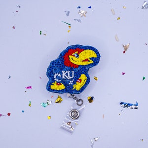May include: A blue and yellow glitter badge reel featuring a cartoon mascot with the letters "KU" on its chest.