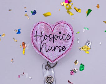 May include: A heart-shaped badge reel with the words "Hospice Nurse" in black script. The badge is pink with a textured appearance and a stitched border. A clear plastic reel and metal clip are attached.