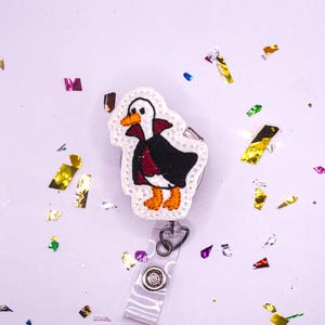 May include: Embroidered badge reel featuring a cartoon duck dressed in a black cape and red vest, with orange feet and beak. The badge reel has a clear plastic strap and a decorative button. The background is white with colorful confetti.