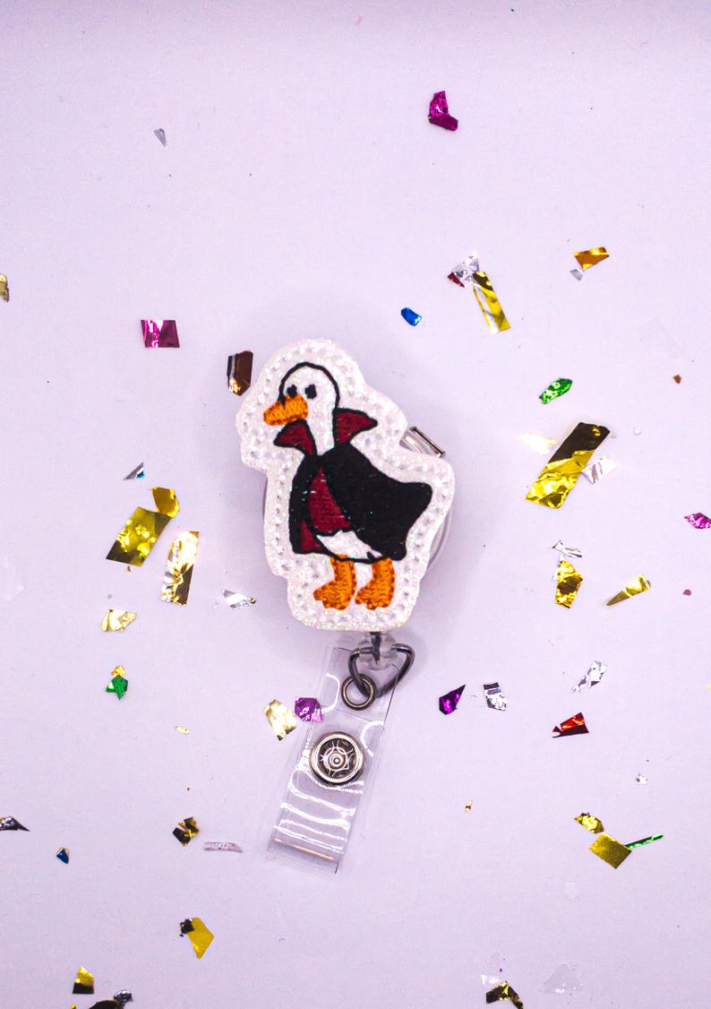 May include: Embroidered badge reel featuring a cartoon duck dressed in a black cape and red vest, with orange feet and beak. The badge reel has a clear plastic strap and a decorative button. The background is white with colorful confetti.