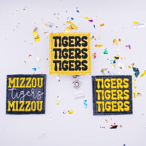 May include: Three glittery badge reels with the words "TIGERS" and "MIZZOU Tigers" in yellow and white lettering on a black background. The reels are square-shaped and surrounded by a black border. Confetti is scattered around the reels.