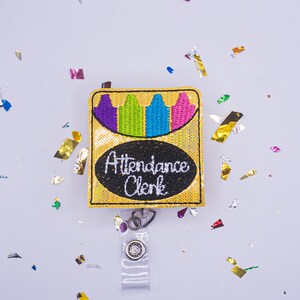May include: A gold sequined badge reel with a crayon box design. The badge features a black oval with the words "Attendance Clerk" in white script. The crayon box has purple, green, blue, and pink crayons. The reel has a clear plastic strap and a metal clip.