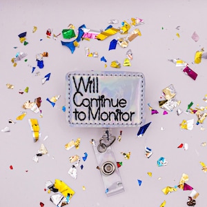 May include: A rectangular badge reel with the text "Will Continue to Monitor" in black lettering. The badge reel has a holographic background and a clear plastic clip. Colorful confetti surrounds the badge reel.