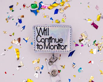 May include: A rectangular badge reel with the text "Will Continue to Monitor" in black lettering. The badge reel has a holographic background and a clear plastic clip. Colorful confetti surrounds the badge reel.