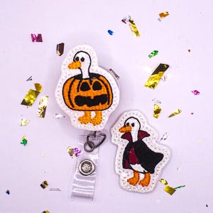 May include: Two embroidered badge reels with duck designs. One is a duck inside an orange pumpkin, and the other is a duck wearing a black cape with a red lining. Both have white borders and are on a white background.
