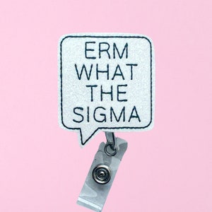 May include: A white badge reel with the text "ERM WHAT THE SIGMA" in black, inside a speech bubble shape. The badge reel has a clear plastic strap and a metal clip, set against a pink background.