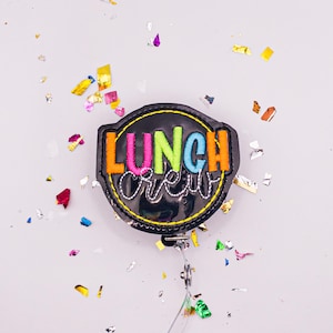 May include: A black badge reel with the words "LUNCH CREW" embroidered in bright colors. The badge reel has a retractable cord and a metal clip. Colorful confetti surrounds the badge reel on a white background.