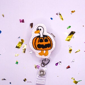 May include: A Halloween-themed badge reel featuring a white duck with an orange pumpkin body and black carved face. The badge reel has a clear plastic strap and a metal clip. Colorful confetti is scattered around the badge reel on a light purple background.
