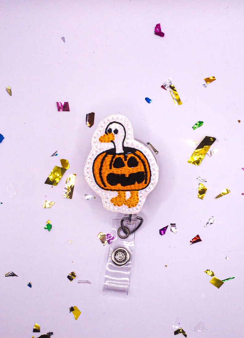 May include: A Halloween-themed badge reel featuring a white duck with an orange pumpkin body and black carved face. The badge reel has a clear plastic strap and a metal clip. Colorful confetti is scattered around the badge reel on a light purple background.