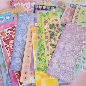 May include: Assortment of colorful sticker sheets with various designs, including hearts, butterflies, musical notes, and bubbles. The stickers are holographic and have a glossy finish.