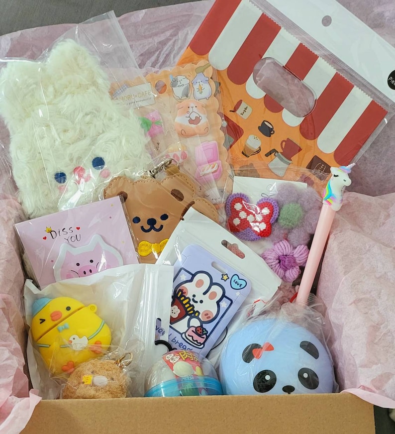 May include: A pink and white gift box filled with various items, including a white fluffy keychain, a brown bear keychain, a purple crocheted heart, a blue panda plush, a yellow chick phone case, and a small pink pig keychain. The box also contains a card that says "Miss You" and a small bag of colorful candies.