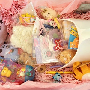 May include: A pink gift box filled with various items, including a white fluffy toy, a small plush animal, a clear plastic container with colorful candies, a book with a blue cover, and a white plastic container with a blue and white design. The box is filled with pink shredded paper.
